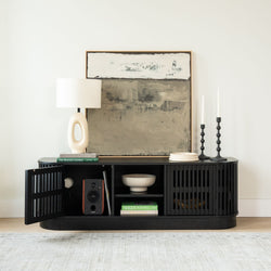 Modern black slatted media console with open shelving styled with abstract beige wall art, oval ceramic table lamp, stacked coffee-table books, white bowl, wood speaker and tall black candlesticks in a minimalist living room