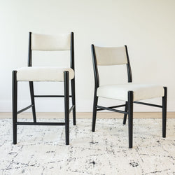 Pair of modern cream upholstered dining chairs with black wooden frames on a patterned cream rug in a minimalist dining room