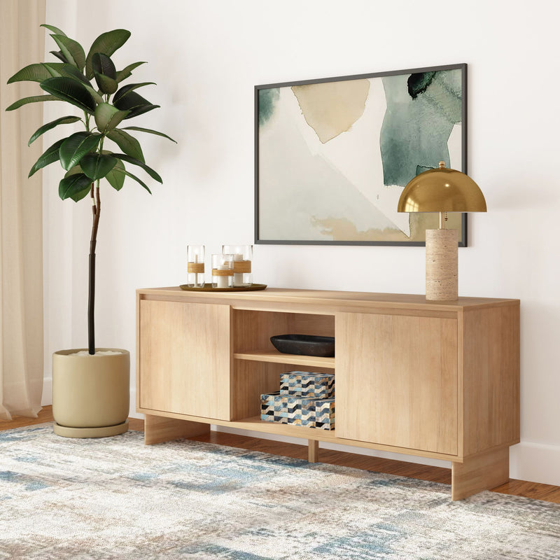Light wood modern sideboard credenza in minimalist living room with gold dome table lamp, abstract green-beige wall art, potted ficus, decorative candles, patterned storage boxes and textured neutral area rug