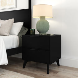 Black modern nightstand with two drawers and mid-century legs topped by a green textured ceramic table lamp with beige shade, scented candle and book beside a black bed on light hardwood floor, contemporary bedroom furniture and décor