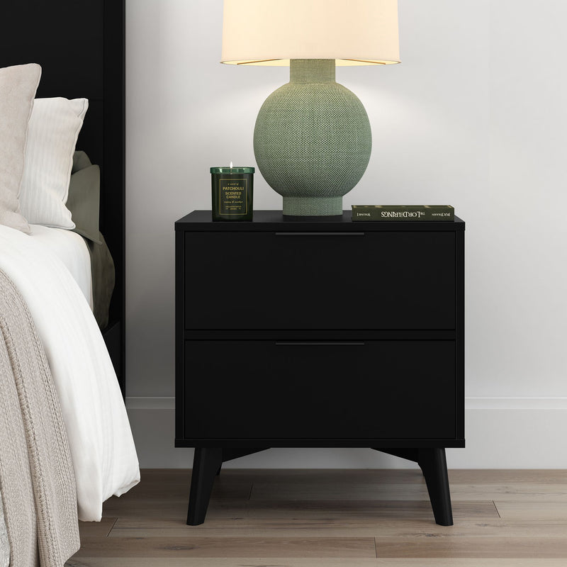 Black mid-century modern two-drawer nightstand beside bed with green textured ceramic lamp, lit patchouli scented candle and hardcover book on light wood floor, contemporary bedroom decor