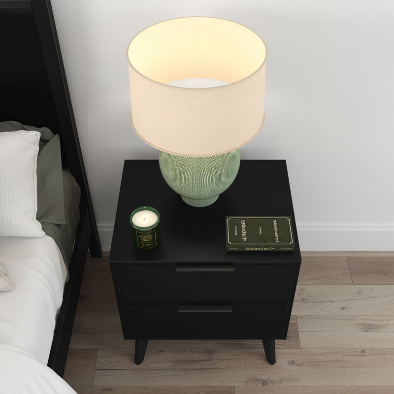 Top-down view of a modern black nightstand with a green textured table lamp and beige shade, scented green candle and book beside a white bed on light hardwood floor — contemporary bedroom decor.