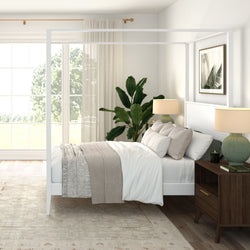 Bright modern bedroom with white four-poster bed, neutral linen bedding and throw, large window with sheer curtains, tall indoor plant, wooden nightstand and green lamp, stylish neutral bedroom decor