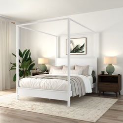 White four-poster canopy bed in a modern minimalist bedroom with neutral bedding, wooden nightstands, green table lamps, potted plant, framed palm artwork, and a textured area rug on hardwood floors.