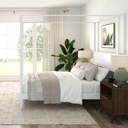 Bright modern farmhouse bedroom with white four-poster canopy bed, neutral layered bedding, large windows, indoor plant and wooden nightstand with lamp