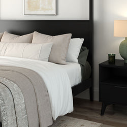 Modern bedroom with black bed frame, white and beige bedding, layered pillows and knit throw, nightstand with lamp and candle