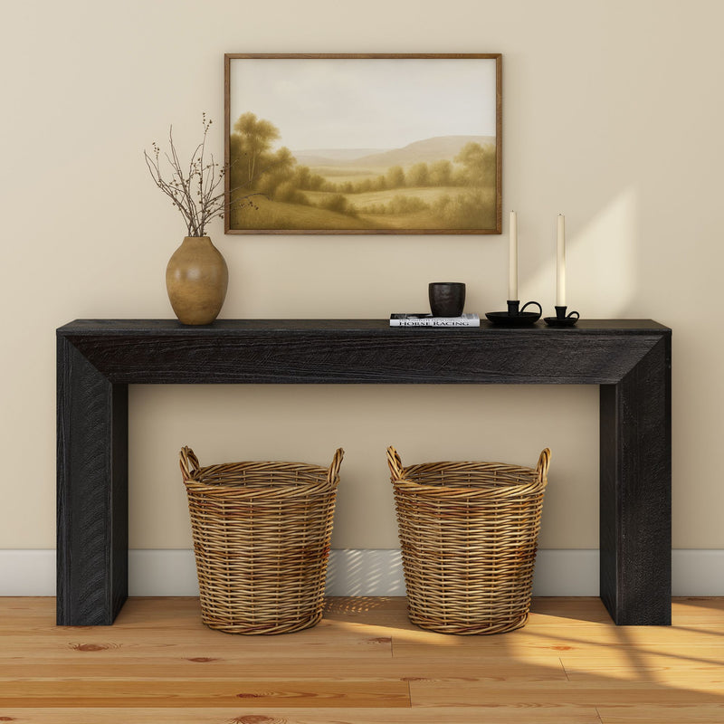 Black wood entryway console table with two woven wicker baskets, ceramic vase with branches, framed landscape print and candles on hardwood floor