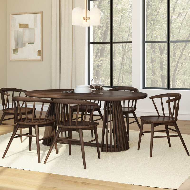 Modern dining room with dark walnut round table and spindle base, six wooden Windsor-style chairs on a neutral rug, pendant light, abstract wall art, and large black-framed windows with tree-lined view, stacked plates and wine glasses