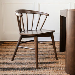 Mid-century modern wooden chair with curved back, placed on striped jute rug in a minimalist interior setting.