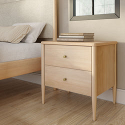 Light wood mid-century modern nightstand with two drawers and brass knobs beside bed on hardwood floor with stacked books on top