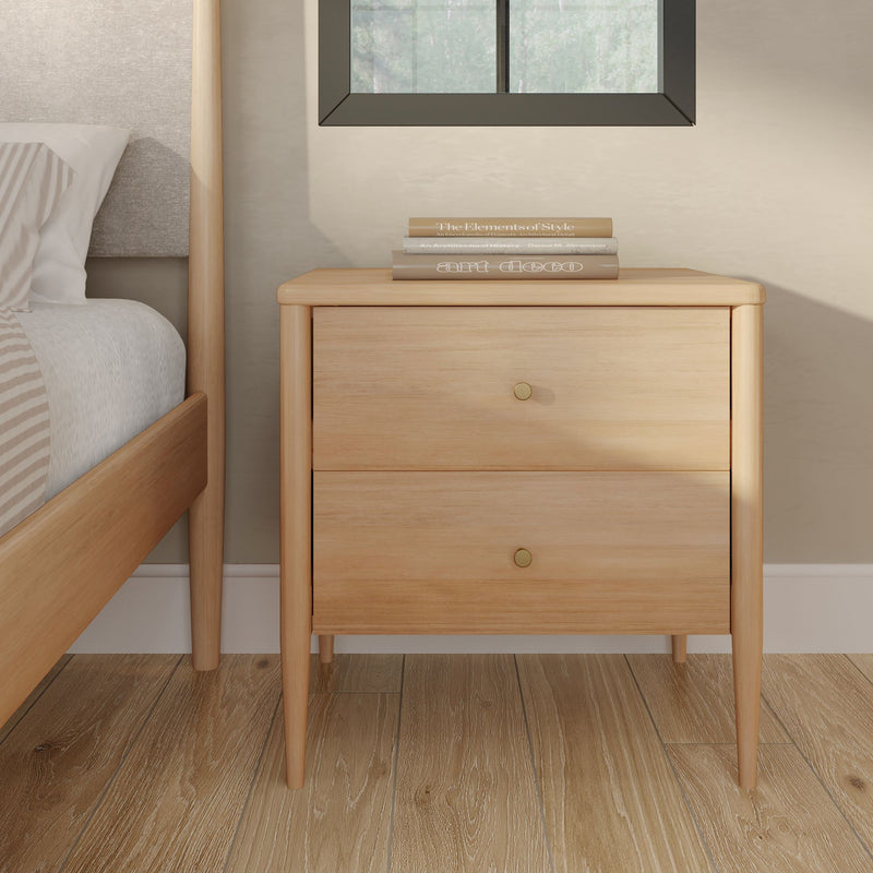 Light wood mid-century modern nightstand, two drawers with brass knobs, stacked books on top next to bed on hardwood floor