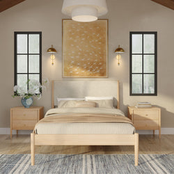 Minimal Scandinavian-style bedroom with a light wood platform bed, striped beige bedding and throw, matching wood nightstands, brass wall sconces, large golden bird artwork above the upholstered headboard, black-framed tall windows, blue patterned rug and a vase of white flowers on the nightstand.