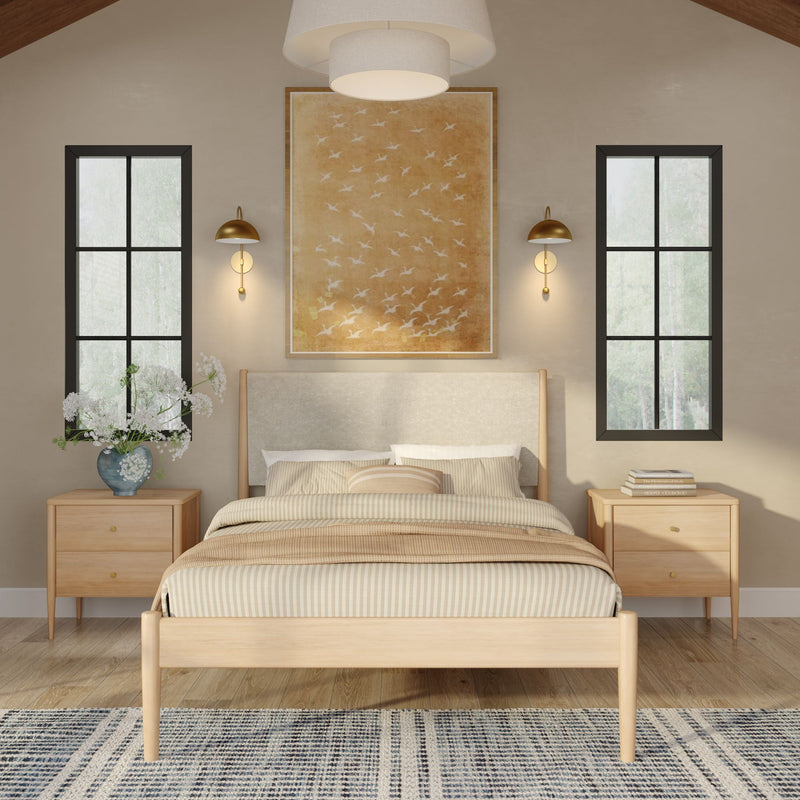 Minimal Scandinavian-style bedroom with a light wood platform bed, striped beige bedding and throw, matching wood nightstands, brass wall sconces, large golden bird artwork above the upholstered headboard, black-framed tall windows, blue patterned rug and a vase of white flowers on the nightstand.