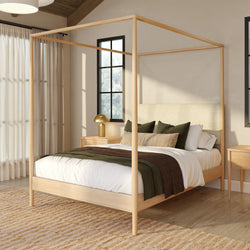 Modern bedroom with light wood four-poster canopy bed, white linens and green-brown bedding, gold bedside lamp, natural light and neutral decor