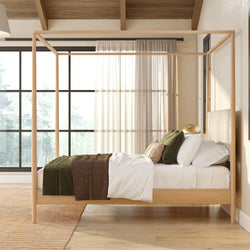 Light-wood canopy bed in a cozy neutral bedroom with large black-framed windows, natural light, green and brown bedding, and hardwood floors