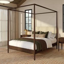 Modern wooden four-poster canopy bed in a neutral farmhouse bedroom with green and brown bedding, wooden nightstands, brass lamp and natural light