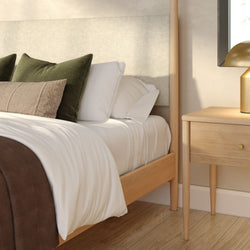 Modern minimalist bedroom with light wood bed frame, crisp white bedding, green textured accent pillows, wooden nightstand and gold table lamp on hardwood floor