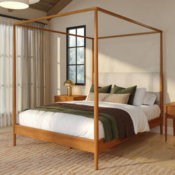 Modern wooden four-poster bed with white linens, green and brown throws, textured beige headboard, wooden nightstands and gold lamp in a sunlit cozy neutral bedroom with hardwood floors and sheer curtains