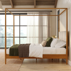 Modern wooden four-poster canopy bed in a minimalist bedroom with white bedding, green and brown throws, textured pillows, large black-framed windows and abundant natural light
