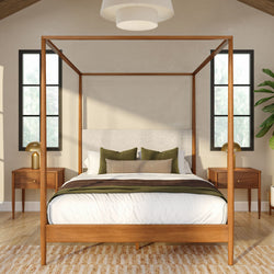 Wooden four-poster bed in a modern neutral bedroom with green accent pillows, matching wooden nightstands, gold table lamps, tall windows, pendant light and indoor plant