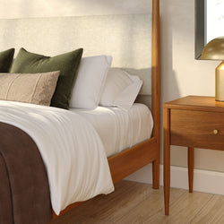 Mid-century modern bedroom corner with wooden bed frame, crisp white bedding, green and beige decorative pillows, wooden nightstand and gold table lamp