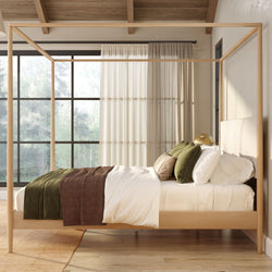 Light-wood four-poster canopy bed in a minimalist rustic bedroom with neutral bedding, green and brown throw pillows, sheer curtains and large window overlooking a forest.