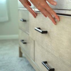 Hands opening a modern wooden dresser drawer with black handles in a bedroom setting.