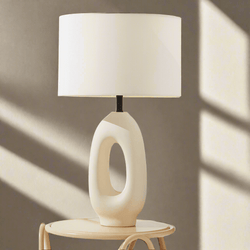 Modern white ceramic table lamp with abstract design on round wooden side table, illuminated by sunlight.