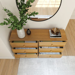 Wooden dresser with open drawers displaying neatly folded clothes, topped with a vase of green foliage, decorative items, and a round mirror reflecting natural light in a modern living room.