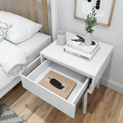 White modern nightstand with open drawer, books, vase with plant, and coffee mug on a wooden floor beside a bed.