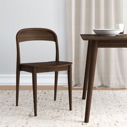 Dark walnut-finish mid-century modern wooden dining chair beside matching table with white bowl, glass, neutral curtains, and textured rug