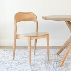 Wooden dining chair next to a round table on a textured rug in a minimalistic room.