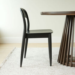 Black wooden dining chair next to round dark‑wood pedestal dining table on light textured rug in minimalist dining room