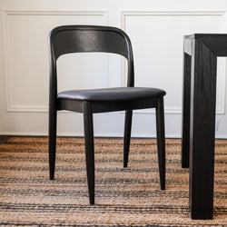 Modern black wooden dining chair with padded seat beside dark wood dining table on striped jute rug in minimalist dining room