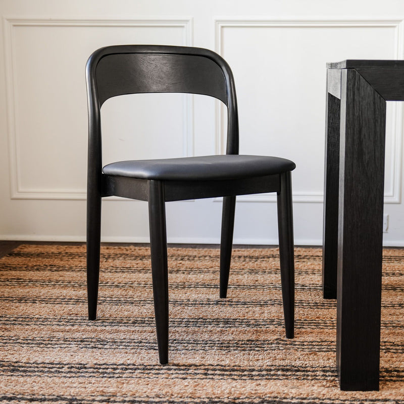 Modern black wooden dining chair with padded seat beside dark wood dining table on striped jute rug in minimalist dining room