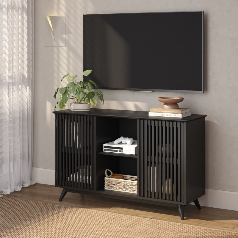 Modern black slatted media console with storage shelves, game console and wicker basket beneath a wall‑mounted flat‑screen TV in a bright living room with potted plant and minimal decor