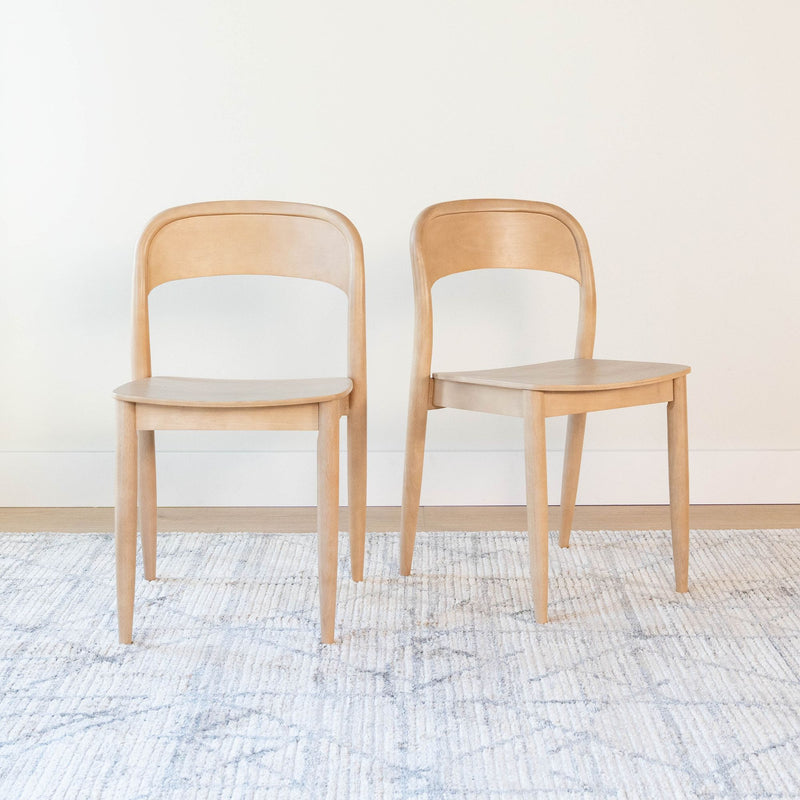 Pair of light wood dining chairs with curved backs in minimalist mid-century modern style on a textured neutral rug against a white wall