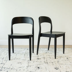 Set of 2 black wooden dining chairs with curved backrests and tapered legs on a textured white area rug in a minimalist dining room