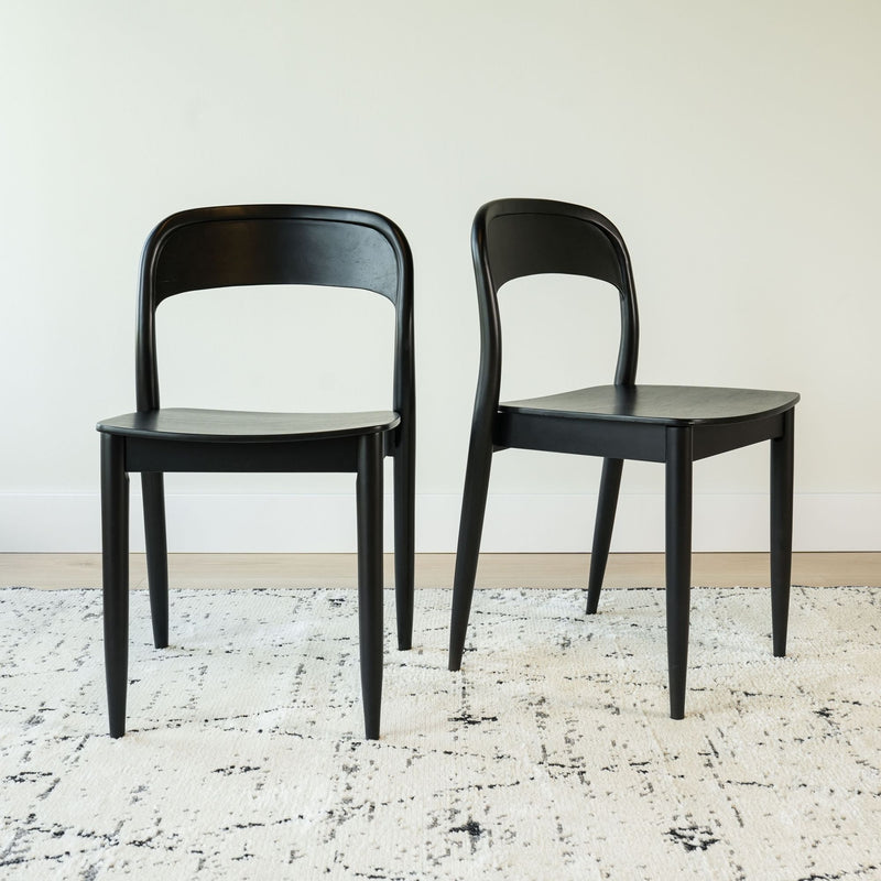 Set of 2 black wooden dining chairs with curved backrests and tapered legs on a textured white area rug in a minimalist dining room