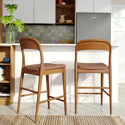 Two mid-century modern wooden bar stools at a white kitchen island with leather seats, green tile backsplash, gray refrigerator and glass vase with plant