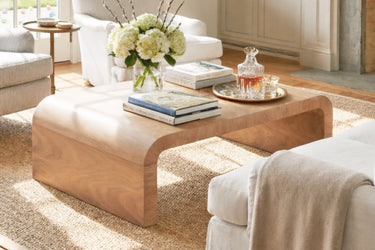 Curved solid-wood coffee table in a neutral living room with linen sofa, jute rug, white hydrangea centerpiece, stacked books and glass decanter tray