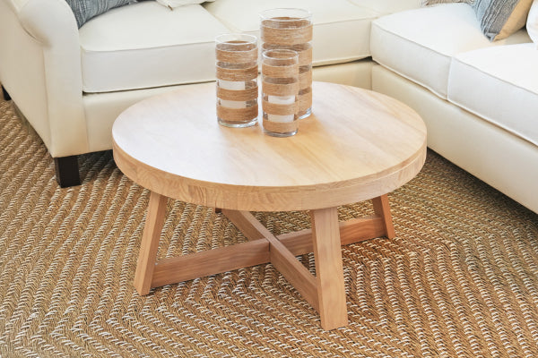 Round natural wood coffee table on woven jute rug between white sectional sofas in bright coastal living room with decorative glass sand-filled jars
