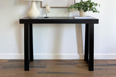 Black modern console table styled as entryway table on hardwood floor with white ceramic lamp, decorative sculpture, potted green plant and book against a white wall