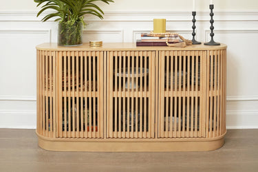 Natural light wood slatted console sideboard cabinet with rounded corners and interior storage, styled with potted plant, books and candles against a white paneled wall