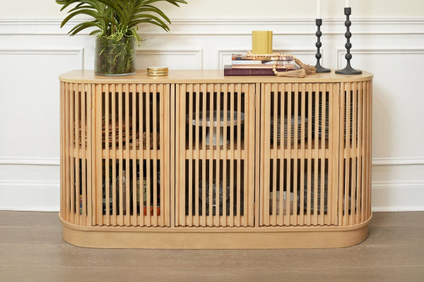Natural light wood slatted console sideboard cabinet with rounded corners and interior storage, styled with potted plant, books and candles against a white paneled wall