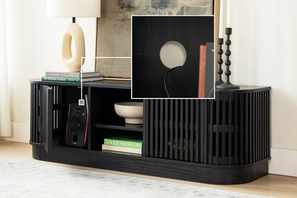 Black wooden slatted media console TV stand with open shelving, built-in cable-management hole, speaker, books and lamp in a modern living room