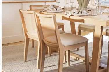 Light natural wood dining table with cane-back chairs and linen seat cushions on a jute rug, set with white plates, cloth napkins and glassware in a bright modern dining room