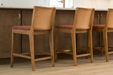 Three wooden bar stools with woven cane backs and rust-colored upholstered seats at a modern kitchen island with white countertop