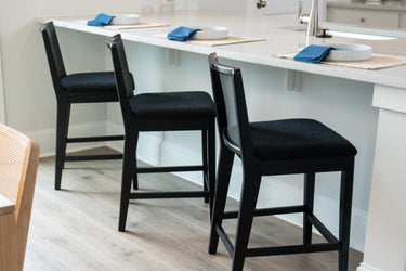 Modern white kitchen island with three black upholstered bar stools, white quartz countertop set with place settings and blue napkins, light wood plank flooring