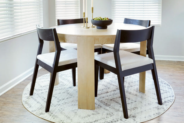 Round light-wood dining table set with four black wood chairs and white cushions on a patterned round rug in a bright modern dining room with window blinds.
